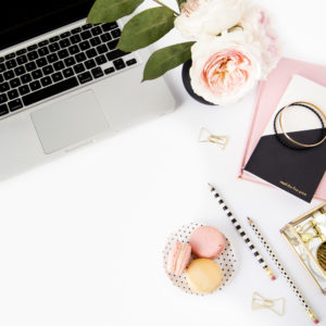 An image of a computer on a desk with macroons to eat, notebooks, flowers, and pencils.