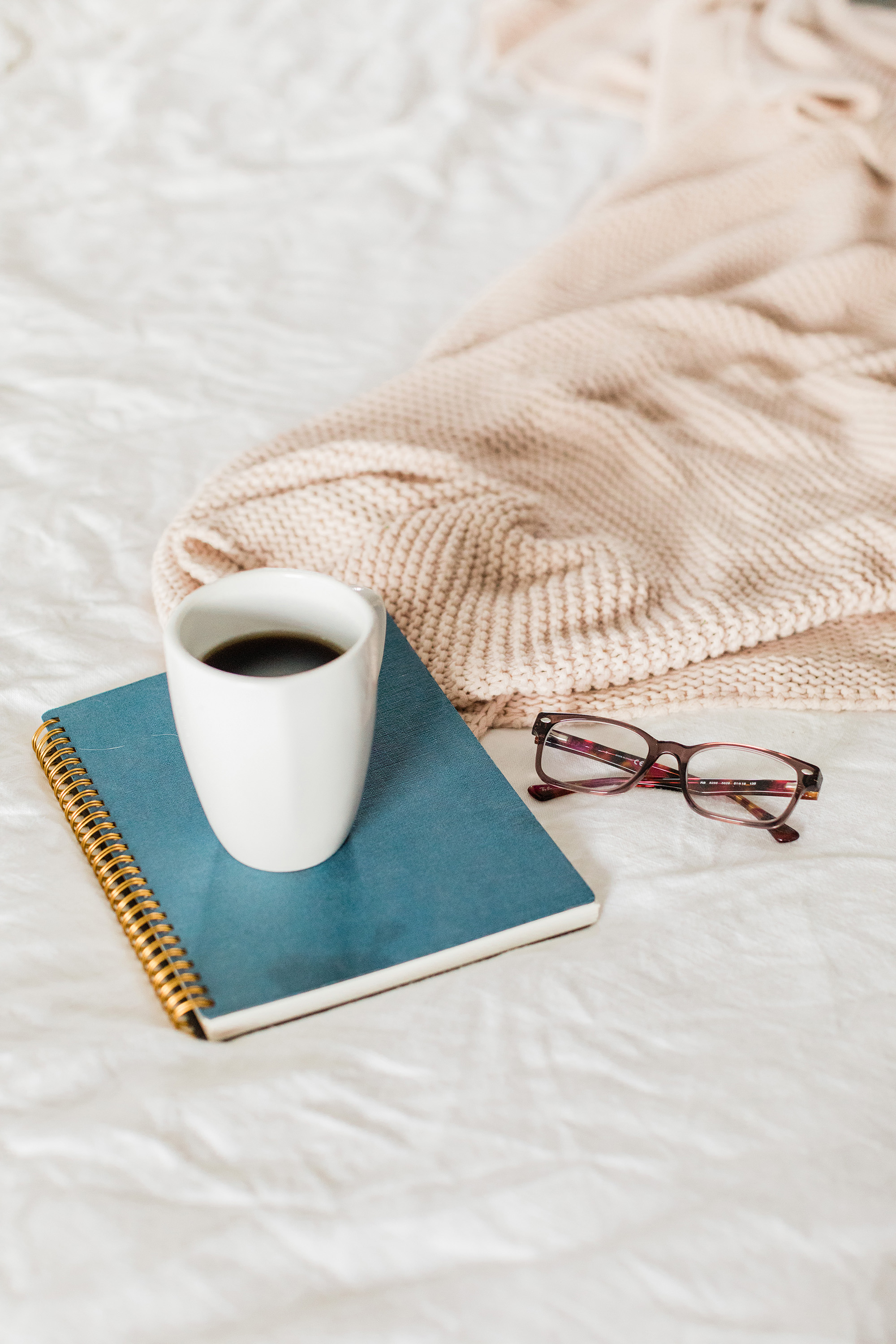 A blue journal on a bed with some coffee on top. Glasses and a blanket on the side.